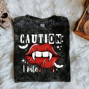 Puede incluir: Camiseta negra con un gráfico de labios de vampiro rojos, colmillos blancos y el texto "CAUTION: I bite." El diseño incluye murciélagos y estrellas blancas. La camiseta está sobre una superficie blanca y esponjosa.