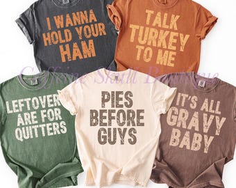 May include: A collection of five short-sleeved t-shirts in various colors, each with a humorous Thanksgiving-themed phrase printed on the front in bold, distressed lettering. The shirts are in shades of gray, orange, green, cream, and brown.