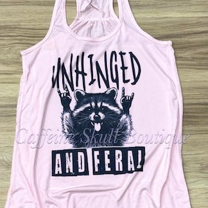 Puede incluir: Camiseta sin mangas rosa claro con un dise&ntilde;o gr&aacute;fico negro de un mapache haciendo el gesto de rock and roll. El texto "UNHINGED" est&aacute; encima del mapache y "AND FERAL" debajo.
