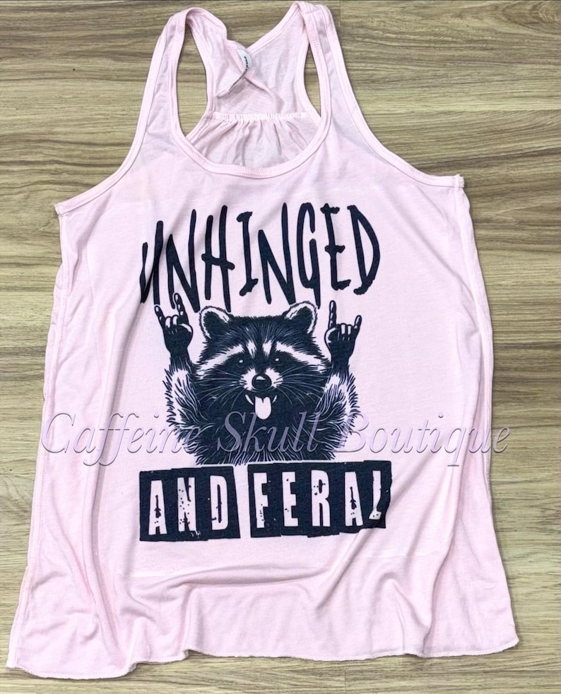 Puede incluir: Camiseta sin mangas rosa claro con un dise&ntilde;o gr&aacute;fico negro de un mapache haciendo el gesto de rock and roll. El texto "UNHINGED" est&aacute; encima del mapache y "AND FERAL" debajo.