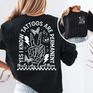 Op de afbeelding: Zwart sweatshirt met een wit grafisch ontwerp met de tekst "Ja, ik weet dat tatoeages permanent zijn, daarom heb ik ze laten zetten". De grafiek bevat een skelet hand die een vredesteken maakt, een bliksem, een schedel en vlinders.