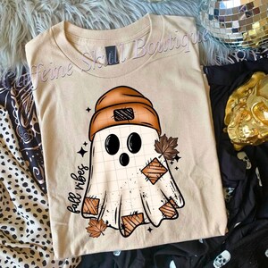 以下が含まれることがあります： ベージュのTシャツ。茶色の帽子とパッチを身に着けた漫画の幽霊が描かれており、秋の葉と「fall vibes」の文字が添えられています。秋にぴったりのデザインです。