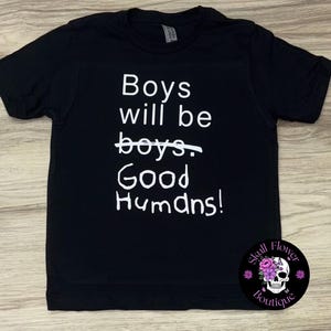 May include: Black t-shirt with white text: "Boys will be boys. Good Humans!" The word "boys" is crossed out. A circular logo with a skull and flowers is in the bottom right corner.