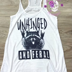 Puede incluir: Camiseta sin mangas blanca con un gr&aacute;fico negro de un mapache haciendo un gesto de rock and roll. El texto "UNHINGED" est&aacute; encima del mapache y "AND FERAL" debajo.
