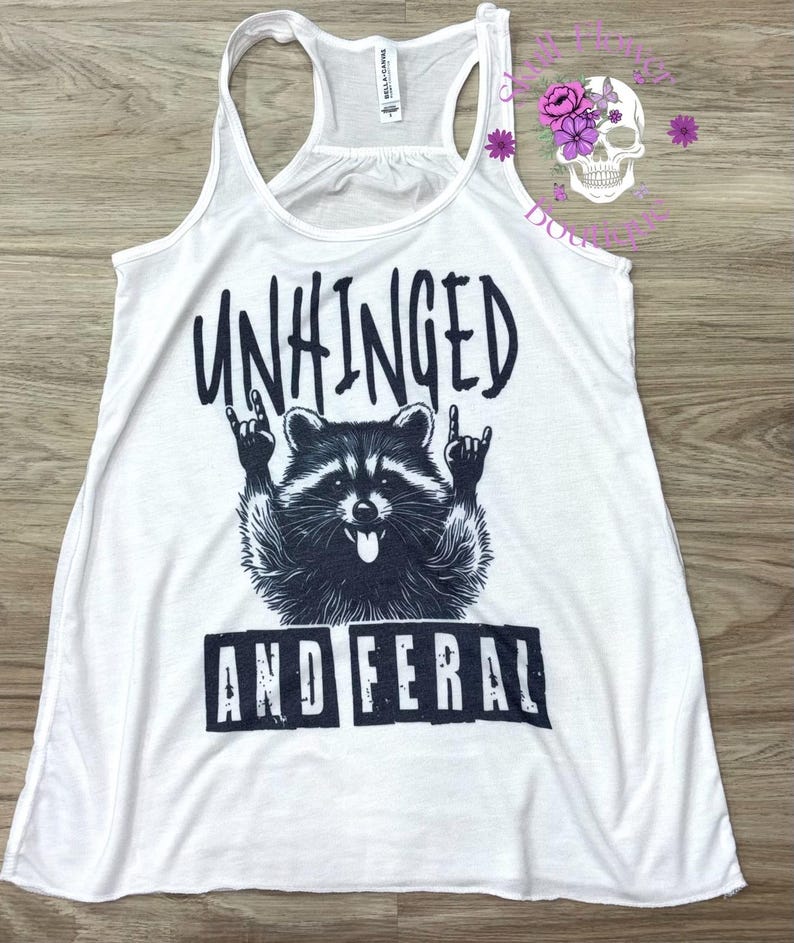 Puede incluir: Camiseta sin mangas blanca con un gr&aacute;fico negro de un mapache haciendo un gesto de rock and roll. El texto "UNHINGED" est&aacute; encima del mapache y "AND FERAL" debajo.