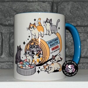 May include: White ceramic mug with a blue handle. The mug features a cartoon illustration of a pill bottle labeled "Serotonin Booster" with a side effect of "Cute Aggression".  The pill bottle is filled with cartoon cats.
