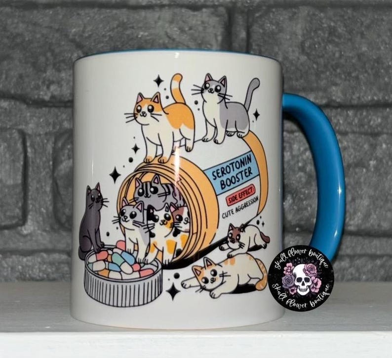 May include: White ceramic mug with a blue handle. The mug features a cartoon illustration of a pill bottle labeled "Serotonin Booster" with a side effect of "Cute Aggression".  The pill bottle is filled with cartoon cats.