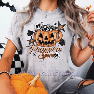 Pode incluir: Camiseta cinza claro com um design de Halloween. O design inclui uma caneca de abóbora com um esqueleto, estrelas e as palavras "Pumpkin Spice". O texto "Caffeine Skull Boutique" também é visível.