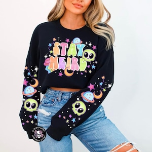 Puede incluir: Sudadera negra recortada con el texto "Stay Weird" en colores arcoíris y gráficos de temática espacial. Las mangas tienen un patrón repetitivo de estrellas, planetas y calaveras.