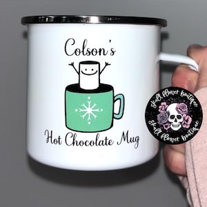 Puede incluir: Taza de esmalte blanco con una ilustración en blanco y negro de un malvavisco sonriente en una taza verde con un diseño de copo de nieve. La taza dice "Colson's Hot Chocolate Mug" y tiene un logotipo en blanco y negro de calavera y flor.
