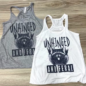 Puede incluir: Dos camisetas sin mangas, una gris y otra blanca, con un gr&aacute;fico negro de un mapache haciendo un gesto de rock and roll con la mano. El texto "UNHINGED AND FERAL" est&aacute; encima y debajo de la ilustraci&oacute;n del mapache.