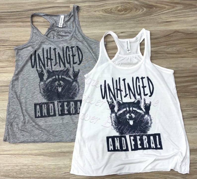 Puede incluir: Dos camisetas sin mangas, una gris y otra blanca, con un gr&aacute;fico negro de un mapache haciendo un gesto de rock and roll con la mano. El texto "UNHINGED AND FERAL" est&aacute; encima y debajo de la ilustraci&oacute;n del mapache.