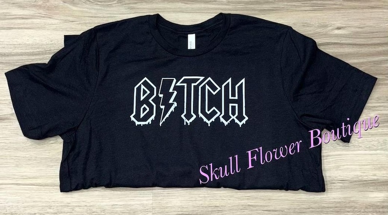 Pode incluir: Camiseta preta com um gr&aacute;fico branco que diz "B*TCH" em uma fonte de metal que pinga.