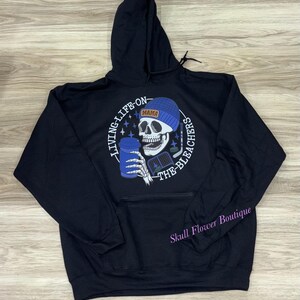 May include: Black hoodie featuring a skull graphic wearing a blue beanie and holding a blue cup and hockey stick. The text reads "LIVING LIFE ON THE BLEACHERS" and "MAMA". The hoodie has a front pocket and a drawstring hood.