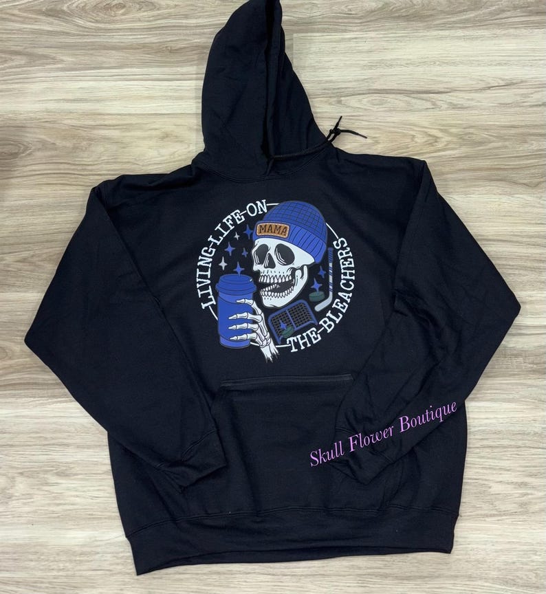 May include: Black hoodie featuring a skull graphic wearing a blue beanie and holding a blue cup and hockey stick. The text reads "LIVING LIFE ON THE BLEACHERS" and "MAMA". The hoodie has a front pocket and a drawstring hood.