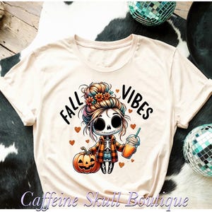 May include: Cream-colored t-shirt with a cartoon skeleton girl design. The design includes the words "FALL VIBES", a jack-o'-lantern, and a beverage. The skeleton girl is wearing a plaid jacket and holding a drink with a straw. The shirt has a round neck and short sleeves.