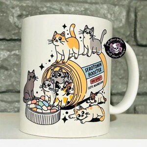 May include: White ceramic mug with a cartoon illustration of cats inside a pill bottle labeled "Serotonin Booster". The text on the pill bottle reads "Side Effect: Cute Aggression".