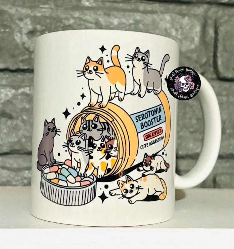 May include: White ceramic mug with a cartoon illustration of cats inside a pill bottle labeled "Serotonin Booster". The text on the pill bottle reads "Side Effect: Cute Aggression".