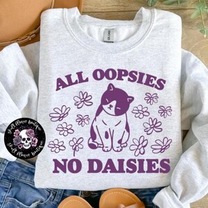 Puede incluir: Una sudadera gris claro con un gr&aacute;fico morado de un gato rodeado de margaritas. El texto "All oopsies no daisies" est&aacute; impreso en morado en la sudadera.