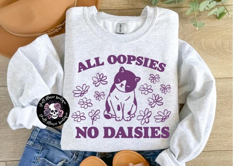 Puede incluir: Una sudadera gris claro con un gr&aacute;fico morado de un gato rodeado de margaritas. El texto "All oopsies no daisies" est&aacute; impreso en morado en la sudadera.