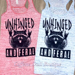 Puede incluir: Dos camisetas sin mangas con espalda de nadador, una roja y otra azul claro, con las palabras "UNHINGED AND FERAL" impresas en negro. Cada camiseta sin mangas presenta un gr&aacute;fico de mapache con las manos en un gesto de rock.