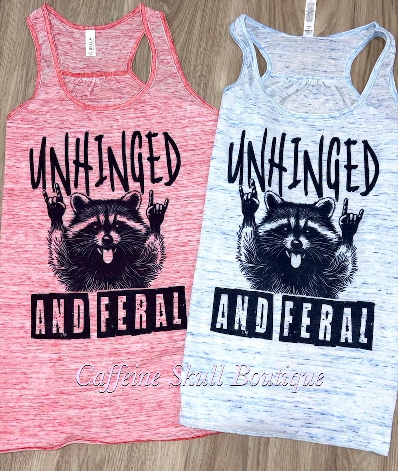 Puede incluir: Dos camisetas sin mangas con espalda de nadador, una roja y otra azul claro, con las palabras "UNHINGED AND FERAL" impresas en negro. Cada camiseta sin mangas presenta un gr&aacute;fico de mapache con las manos en un gesto de rock.