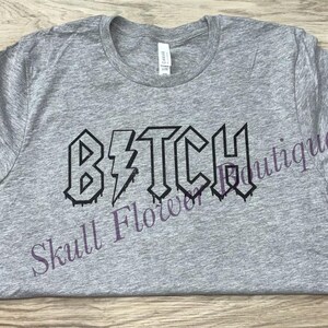 Pode incluir: Camiseta cinza com a palavra "B*TCH" impressa em preto, com um raio no meio da palavra. As letras s&atilde;o estilizadas para parecer um logotipo de banda de rock.