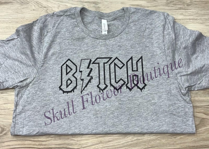 Pode incluir: Camiseta cinza com a palavra "B*TCH" impressa em preto, com um raio no meio da palavra. As letras s&atilde;o estilizadas para parecer um logotipo de banda de rock.