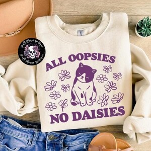 Puede incluir: Una sudadera de color crema con un gr&aacute;fico morado de un gato con el texto "All oopsies no daisies".