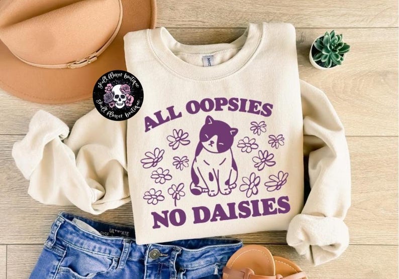 Puede incluir: Una sudadera de color crema con un gr&aacute;fico morado de un gato con el texto "All oopsies no daisies".