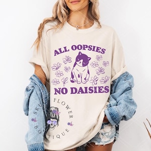 Puede incluir: Una camiseta blanca con el texto "All oopsies no daisies" en letras moradas. Una ilustraci&oacute;n de un gato morado y blanco est&aacute; en el centro del texto. El texto "Flower Sku Boutique" est&aacute; en letras moradas en la parte inferior de la camiseta.