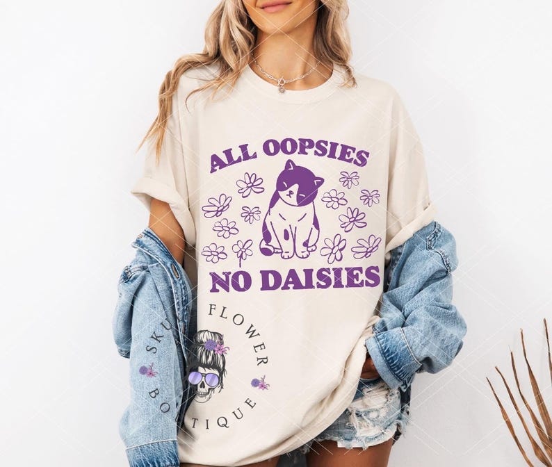 Puede incluir: Una camiseta blanca con el texto "All oopsies no daisies" en letras moradas. Una ilustraci&oacute;n de un gato morado y blanco est&aacute; en el centro del texto. El texto "Flower Sku Boutique" est&aacute; en letras moradas en la parte inferior de la camiseta.
