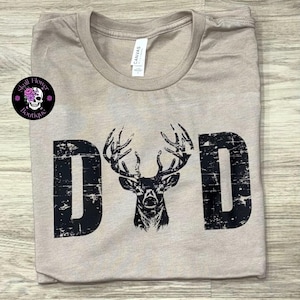 Puede incluir: Camiseta beige con la palabra "DAD" en negro desgastado y un dibujo de una cabeza de ciervo. La camiseta tiene cuello redondo y una etiqueta "CANVAS".