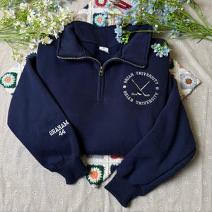 Op de afbeelding: Marineblauw sweatshirt met een kwartrits en lange mouwen. De linkermouw heeft de witte borduursel "GRAHAM 44". De borst heeft een wit borduurontwerp met de woorden "BRIAR UNIVERSITY" en gekruiste hockeysticks. Het sweatshirt is een cropped stijl.