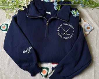 May include: Navy blue sweatshirt with a quarter-zip closure and long sleeves. The left sleeve has "GRAHAM 44" embroidered in white. The chest features a white embroidered design with the words "BRIAR UNIVERSITY" and crossed hockey sticks. The sweatshirt is a cropped style.