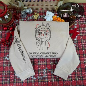 Puede incluir: Sudadera beige con un diseño gráfico que presenta una cara con detalles rojos y el texto "I'M SO MUCH MORE THAN WHAT YOU MADE ME". La manga izquierda tiene un diseño de espada y hojas.
