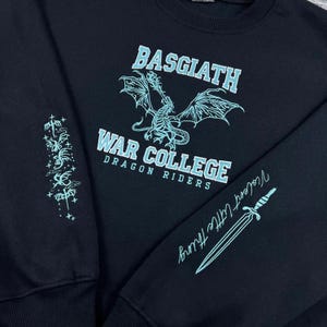 May include: Black sweatshirt with the text "BASGIATH WAR COLLEGE DRAGON RIDERS" in light blue. A dragon graphic is centered above the text. The sleeve has a design and the words "Violent Little Thing" with a sword graphic.
