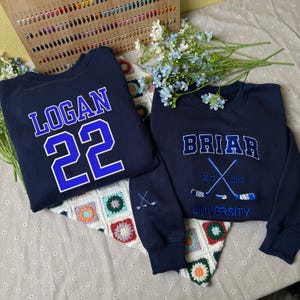 Könnte beinhalten: Zwei marineblaue Sweatshirts. Eines zeigt "LOGAN" und die Nummer "22" in Weiß und Blau. Das andere hat "BRIAR" über gekreuzten Hockeyschlägern und "UNIVERSITY" darunter. Beide haben Hockeyschläger-Grafiken auf den Ärmeln.
