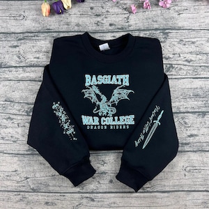 May include: Black sweatshirt with the text "BASGIATH WAR COLLEGE DRAGON RIDERS" in a light blue font. A dragon graphic is centered above the text. The left sleeve has a light blue design, and the right sleeve has a sword graphic and the text "Violent Little Thing".