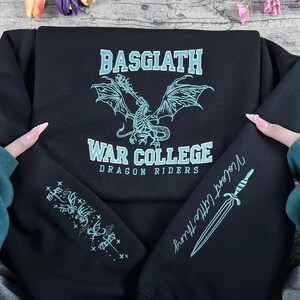 May include: Black sweatshirt with "BASGIATH WAR COLLEGE DRAGON RIDERS" text and a dragon graphic in light blue. The left sleeve has a floral design, and the right sleeve has a dagger and the text "Violent Little Thing".