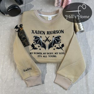 Op de afbeelding: Beige sweatshirt met de tekst "XADEN RIORSON" en "MY POWER, MY BODY, MY SOUL IT'S ALL YOURS." met een ontwerp van gekruiste zwaarden en vleermuisvleugels. De mouw leest "Xaddy". De sweatshirt heeft een geribbelde kraag en manchetten.