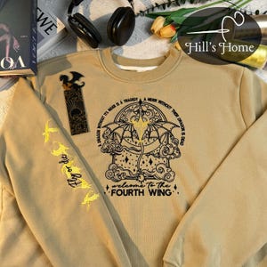 May include: A tan sweatshirt with a black embroidered design of a dragon and clouds. The text "A dragon without it's rider is a tragedy. A rider without their dragon is dead. Welcome to the Fourth Wing" is also embroidered on the sweatshirt.