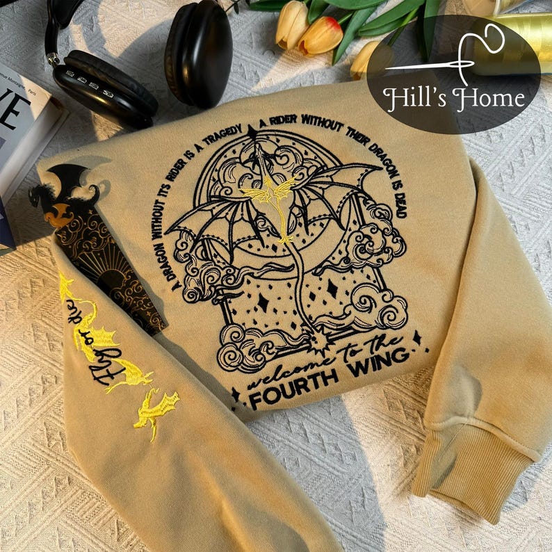 May include: A tan sweatshirt with a black embroidered design of a dragon and clouds. The text "A dragon without its rider is a tragedy. A rider without their dragon is dead. Welcome to the Fourth Wing." is embroidered in black on the sweatshirt.
