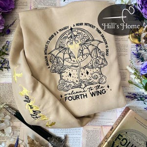 May include: A tan sweatshirt with a black embroidered design of a dragon and clouds. The text "A dragon without its rider is a tragedy. A rider without their dragon is dead. Welcome to the Fourth Wing" is embroidered on the sweatshirt.