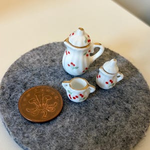 Può includere: Servizio da tè in miniatura in ceramica bianca con dettagli di ciliegie rosse e finiture dorate. Il set comprende una teiera, una zuccheriera e una lattiera. Una moneta britannica da due pence è accanto per la scala. Il set è su un cerchio di feltro grigio.