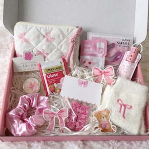 May include: A pink gift box filled with various items, including a white quilted cosmetic bag with pink bow accents, face masks, a scrunchie, a candle, tea, and a small teddy bear sticker. The box is lined with pink paper and sits on a white, fluffy surface.