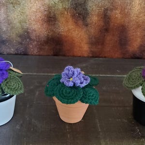Könnte beinhalten: Drei gehäkelte Afrikanische Veilchen Pflanzen. Jede Pflanze hat eine leuchtend violette Blüte mit gelbem Zentrum, umgeben von grünen Blättern. Die Pflanzen befinden sich in verschiedenen Töpfen: weiß, Terrakotta und schwarz.