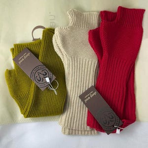 May include: Three pairs of ribbed knit fingerless gloves in olive green, beige, and red. Each pair has a tag that reads "sheep wool quality". The gloves are displayed on a light-colored surface.