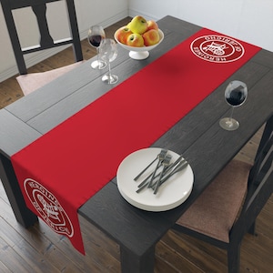 May include: Red table runner with a white circle design featuring the text "HEROINES OF DETROIT" and a graphic of a train engine. The runner is on a dark wood table with a plate of silverware and a glass of wine.