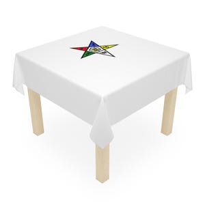 May include: A white square tablecloth with a colorful star emblem, set on a table with light-colored wooden legs. The star features red, blue, yellow, green, and white points, with various symbols within.